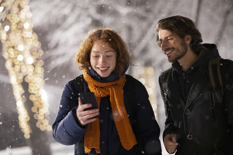Two people smiling as they look at a smartphone outdoors on a snowy winter evening, surrounded by warm Christmas lights and a festive holiday atmosphere
