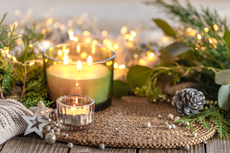 A lit candle surrounded by evergreen branches, fairy lights, and festive ornaments on a woven mat, creating a warm and cozy Christmas atmosphere.