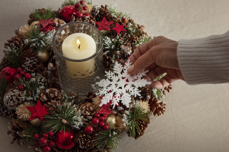 A hand placing a white snowflake ornament onto a festive Christmas wreath decorated with pinecones, evergreen branches, red accents, and a lit candle at the center
