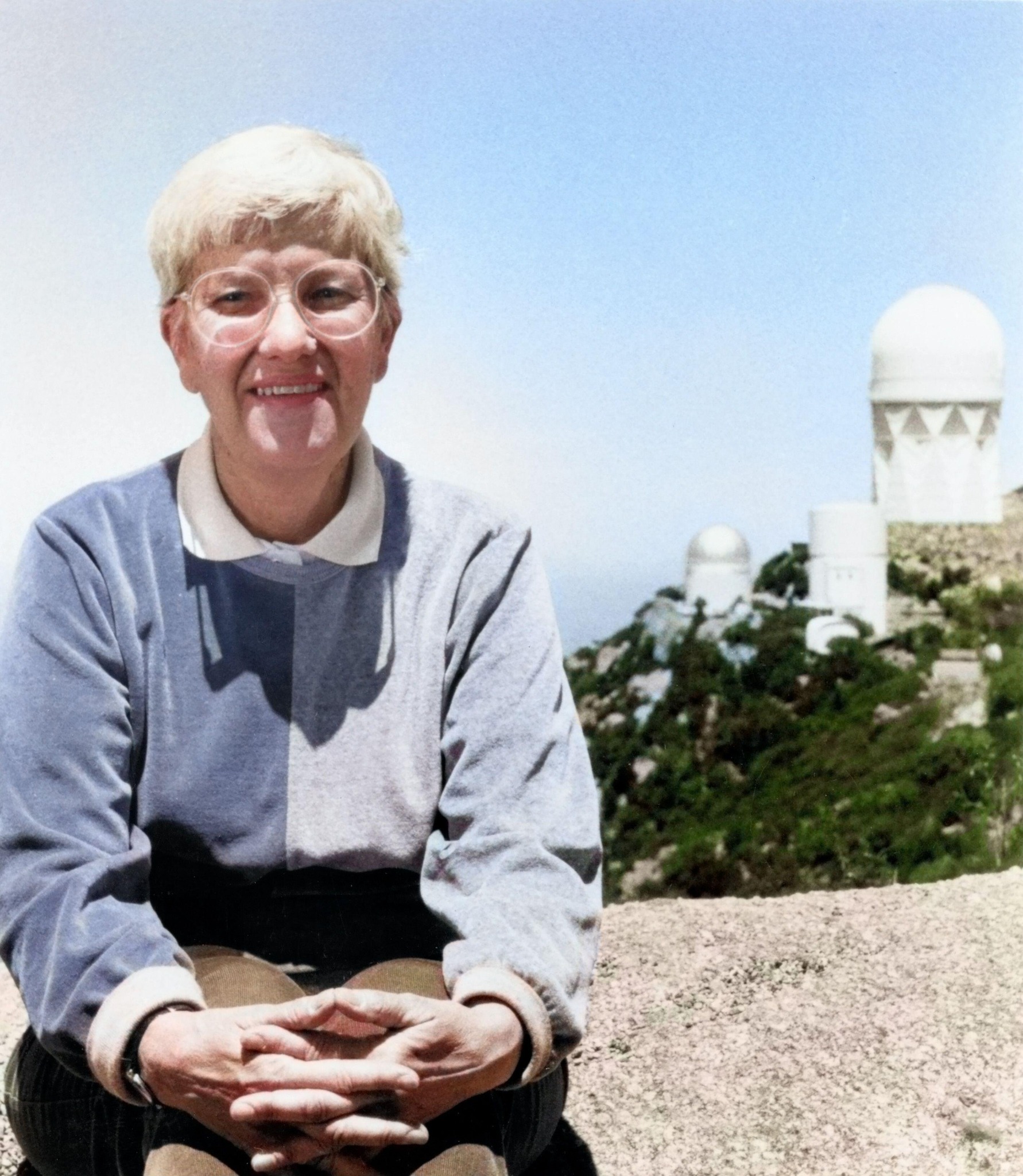 Vera Rubin smiles while seated on a stone ledge outdoors, hands clasped, with white astronomical observatory domes rising on a hillside behind her beneath a clear blue sky.