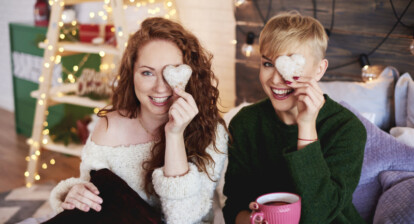 Two women sitting together in a cozy, Christmas-decorated room, smiling while holding heart-shaped gingerbread cookies over one eye