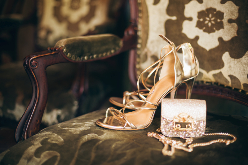 Elegant gold high-heeled sandals and a sparkling clutch purse placed on a vintage upholstered chair, creating a luxurious fashion scene; Source: https://www.freepik.com/free-photo/luxury-fashion-woman-accessories-golden-heeled-shoes-little-evening-purse-elegant-style-vintage-style-sandals-footwear_12152478.htm
