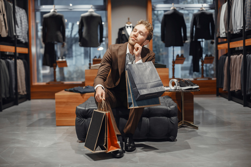 Well-dressed man sitting in a luxury clothing boutique holding shopping bags, surrounded by elegant suits and high-end fashion displays, symbolizing premium brand shopping. Source: https://www.freepik.com/free-photo/sale-shopping-fashion-style-people-concept-elegant-young-man-choosing-footwear-mall_17091285.htm