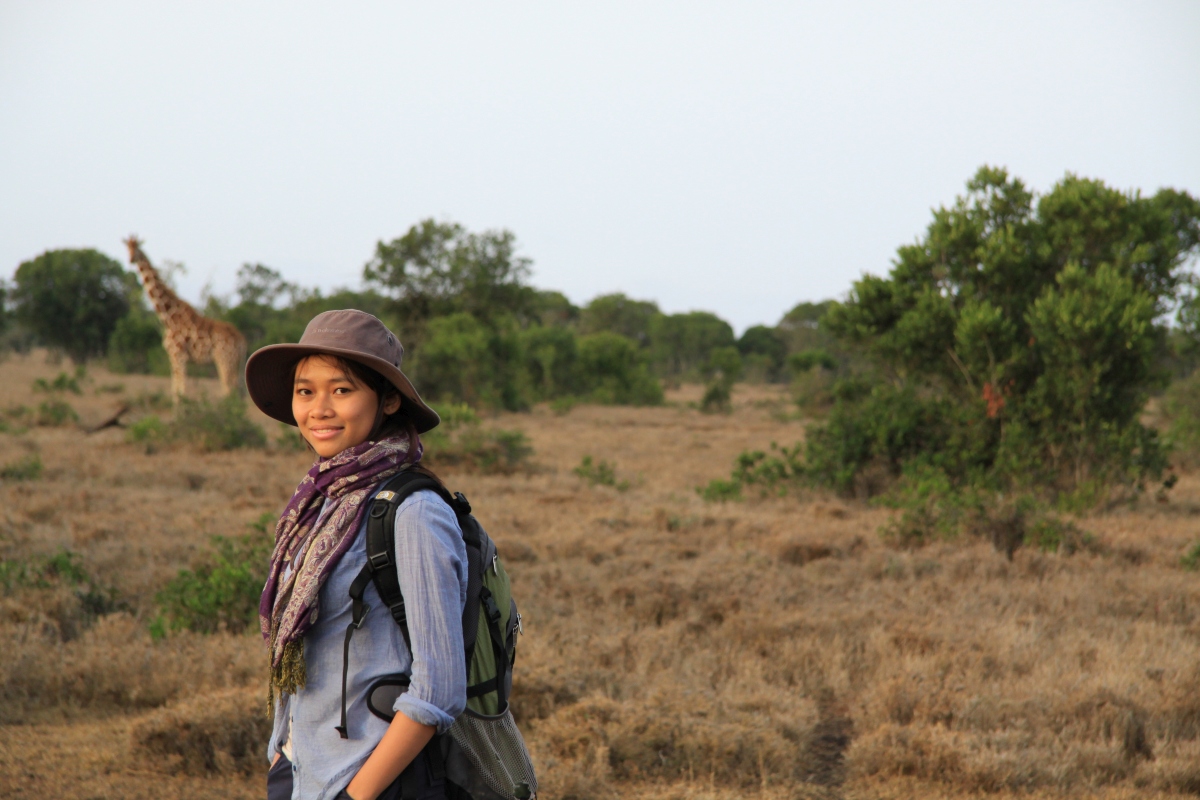 Trang Nguyen standing in a wildlife reserve in Kenya with a giraffe in the background, representing her conservation work in the field. 