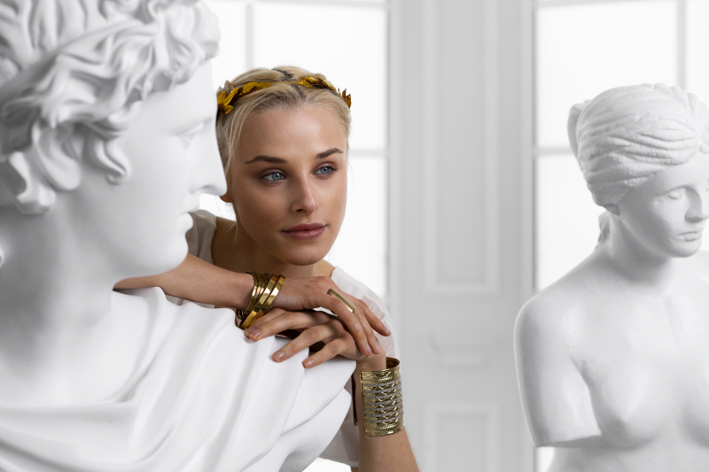 Woman styled as a classical Greek figure, wearing a gold headpiece and jewelry, leaning thoughtfully beside white marble sculptures in a bright studio. Source: https://www.freepik.com/free-photo/front-view-woman-posing-as-greek-goddess_37006330.htm#