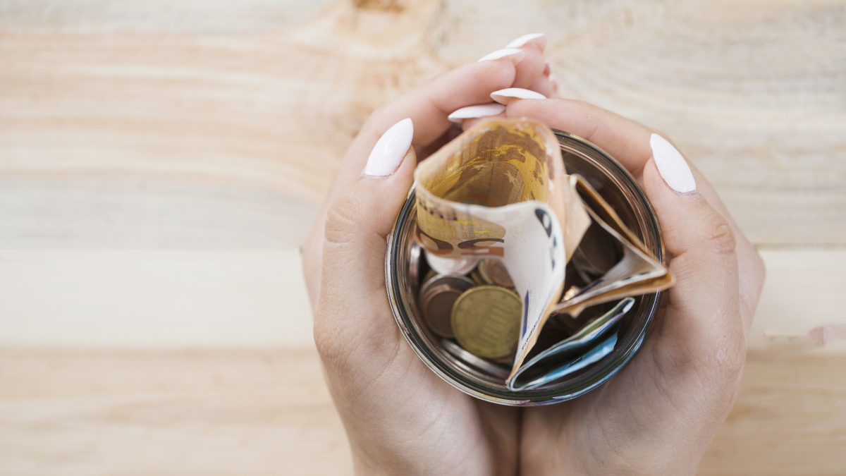 Hands holding a glass jar filled with coins and folded banknotes, symbolizing savings or extra income., Source: https://www.freepik.com/free-photo/woman-s-hand-holding-glass-jar-with-euro-notes-coins-wooden-backdrop_3088564.htm 