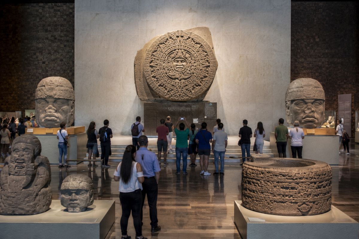 Visitors viewing the Aztec Sun Stone, Olmec colossal heads, and ancient Mesoamerican artifacts inside a museum gallery dedicated to pre-Columbian civilizations. Image created with AI