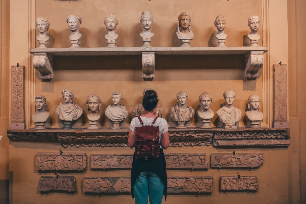 Museum visitor viewing a collection of ancient marble busts and archaeological artifacts displayed on a gallery wall. Source: https://www.magnific.com/free-photo/renaissance-architecture-street-lamp-louvre-museum-paris_4438345.htm