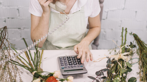 Small business owner taking a phone call while calculating expenses at her flower workshop. Source: https://www.magnific.com/free-photo/crop-florist-using-calculator-phone-conversation_1759217.htm