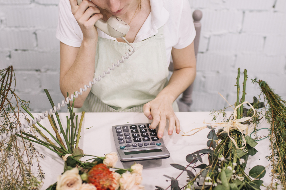 Small business owner taking a phone call while calculating expenses at her flower workshop. Source: https://www.magnific.com/free-photo/crop-florist-using-calculator-phone-conversation_1759217.htm