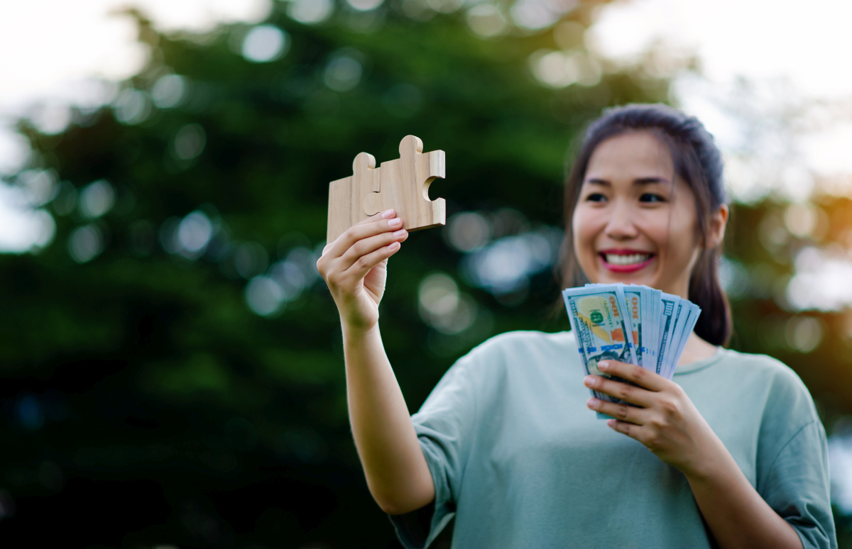 Woman holding banknotes and wooden puzzle pieces, symbolising the evaluation of profitable roles within a portfolio career. Source: https://www.magnific.com/premium-photo/dollar-hands-asian-woman-savings-investments-salary-income-cash-flow-happiness-financial-success-investments_65046614.htm 
