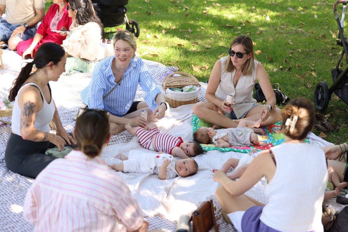 Mothers sit together with their babies on picnic blankets during a Villagehood gathering in a park.Caption: Mothers and babies connect at a Villagehood community picnic. Photo credit: Photo courtesy of Brittany Bloomer / Villagehood.