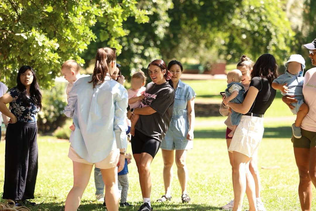 A diverse group of mothers talk and hold their babies at a Villagehood community gathering in a park.Caption: Villagehood brings local mothers together to build friendships and share motherhood in real life. Photo credit: Photo courtesy of Brittany Bloomer / Villagehood.