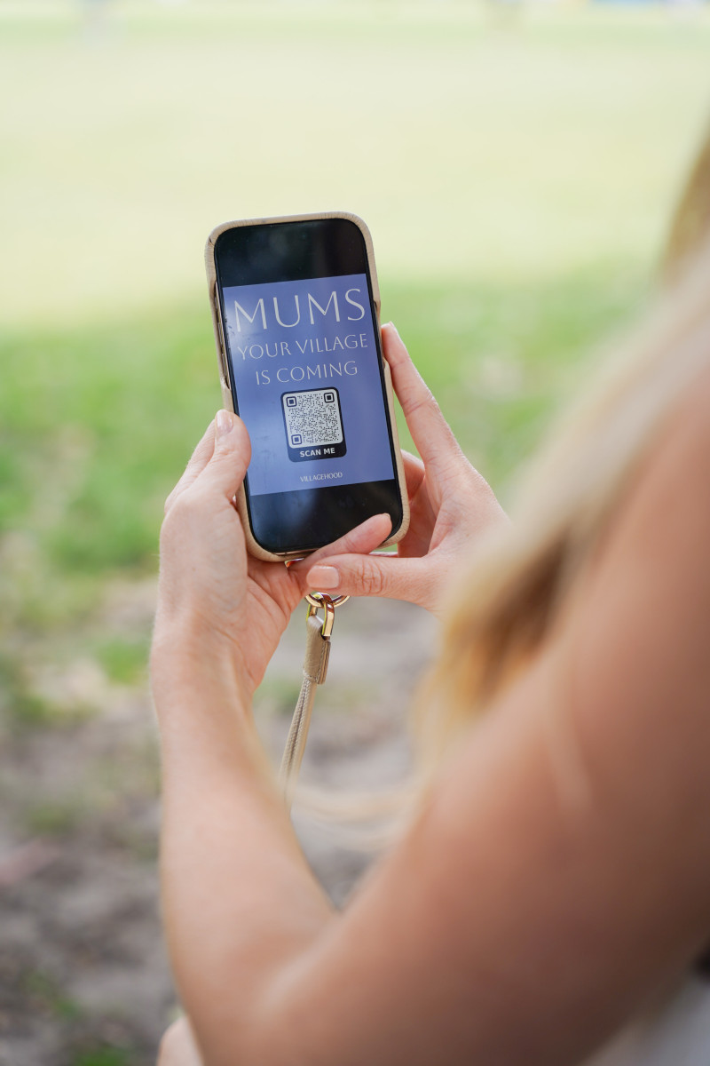A woman holds a phone displaying a Villagehood QR code and the message “Mums, your village is coming.”Caption: Villagehood uses technology to help mothers find connection and community in real life. Photo credit: Photo courtesy of Brittany Bloomer / Villagehood.